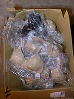 Box filled with many unopened bras, mostly nude and black, in original packaging inside a cardboard box.