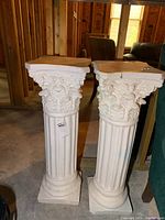 Two white ceramic Corinthian-style columns standing side by side. Both have fluted shafts and detailed capitals with acanthus leaf motifs. Square bases and flat tops are visible.