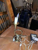 View of brass trumpet-shaped electric lamp on table showing overall structure, coiled tubing, circular base, white electrical cord and switch.
