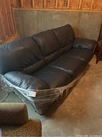 Black leather full-size sofa with three seat and back cushions, shown in corner of unfinished basement with concrete floor and walls. Sofa partially wrapped in plastic film around base.