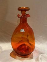 Full view of orange Blenko glass decanter with label and stopper.