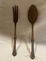 Two serving utensils: one fork and one spoon with teak wood functional ends and silver handles on a light background.