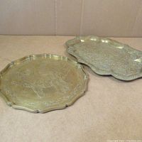 Photo showing two brass trays placed on a cardboard surface, displaying their overall shape and engraved design features.