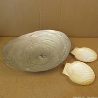 Full view of the large metal seashell dish with hammered texture and two smaller seashells inside, next to two natural real seashells.