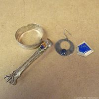 Four assorted sterling silver items on a beige surface: cuff bracelet, sugar tongs with enamel, hoop earring with stone, shield pin with blue stone.