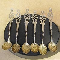 Set of six small silver spoons showing shell-shaped bowls with gold tone and intricate openwork handles with a twisted shaft design, laid out on a black display stand.