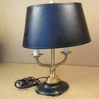 Front view of brass desk lamp showing black lampshade, two candle-style bulb holders, and round black and brass base with power cord coiled beside it.