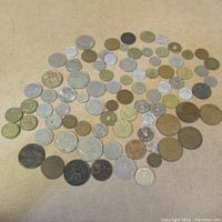 Wide shot of 63 assorted vintage and antique world coins of various sizes and metals spread on a surface, showing wear from age and use.