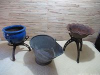 Photo of blue ceramic flower pot on metal plant stand, metal pail, and metal bowl on metal stand on a cream colored textured surface with a wood textured background.
