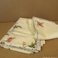 Folded cream-colored tablecloth and napkins showing floral petit-point embroidery and blue border.