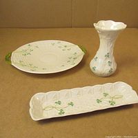 Three white Belleek porcelain items: round platter with green handles and shamrock decoration, rectangular shamrock tray, and shamrock vase with flared rim.