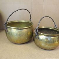 Two antique brass pots with rounded bodies and attached black metal handles, standing side by side on cardboard.