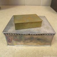 Two small vintage trinket boxes stacked, larger silver-toned with decorative edge and smaller brass-toned on top.