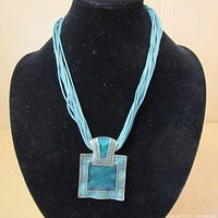 Turquoise multi-strand beaded necklace with large square blue-green enamel pendant on black velvet bust