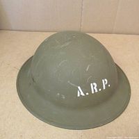 Front angled view of an olive-green steel helmet with white "A.R.P." stenciling, showing surface scratches and wear.