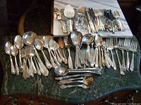 Complete set of silver-plated knives, forks, spoons and serving pieces arranged on tables, showing varied patterns and styles including some acrylic handled pieces.