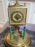 Front angle of old German anniversary clock showing square face with Arabic numerals, brass details, twisted green columns, and green pendulum balls on brass base