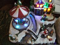 Photo showing the carousel scene with lit horses, house with multi-colored lights, Santa figure, and snow-covered platform with bridges and stream.