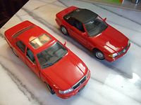 Photo of two red diecast model cars placed on a white marble surface, showing Lexus LS400 and Mercedes Benz 500SL models side by side.