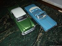 Two vintage model cars on a green marble surface: left green and white 1953 Chevy Belair Custom, right blue 1962 Plymouth Fury convertible.