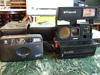 Two vintage cameras on a green marble surface: Minolta AF 10 Freedom 35mm camera and Polaroid Sun 660 instant camera, both with carrying cases visible behind.