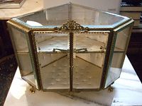 Front angled view of hexagonal brass and beveled glass display cabinet with tufted silk interior showing intricate brass detailing and claw feet.