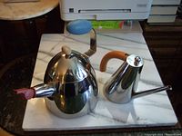 Front view showing both kettles together on a marble surface; Alessi whistle kettle showing blue handle and red whistle next to Giannini kettle with wooden handle