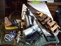 Overview photo showing 3 jig saws with cords, 1 glue gun, some hand saws with wooden handles, electric drill, and other tools on a marble table.