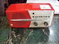 Front left angled view showing full radio unit, tuning scale, black knobs, and red casing on green marble surface.