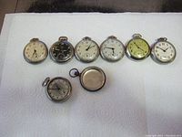 Lot overview showing all 8 vintage pocket watches on white towel background, various dials and cases, some bezels cracked or missing.