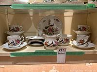 Display shelf with full china set including six cups and saucers, side plates, serving bowl, serving dish, and creamer pitcher all decorated with peacock and floral patterns, some wear visible.