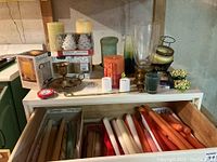 Photo showing assortment of wax candles placed on top of table and inside drawer, plus brass and glass candle holders.