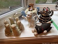 View of all items on windowsill including cat figurines arranged around votive holder, teddy bear teapot with clock, white cat teapot, black-striped cat teapot, and stuffed cat figure.