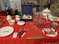 Christmas-themed dishes and assorted holiday dishware displayed on red tablecloth. Includes plates, teapots, cream and sugar set, glass cups, serving platters, and small decorative items.