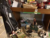 Golf bag with clubs, shoes, books, balls, figurines and other accessories on and around a wooden shelf in a basement setting