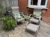 Two patio chairs with matching ottoman, each chair has a tropical leaf throw cushion, next to a hanging plant stand with fern
