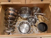 Top view of four Lagostina stainless steel pots and one Calphalon non-stick pot with several glass lids organized in a drawer.
