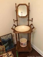 Full view of the wooden washstand with porcelain basin and floral pitcher on lower shelf, placed beside wall with some framed artwork visible on floor.