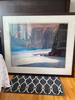 Framed print showing a coastal beach landscape with cliffs and sand, placed on floor with window reflection on glass.
