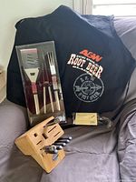Wooden knife block with knives, BBQ apron and packaged BBQ tools on a gray surface with window background.