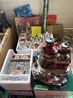 Photo of five plug-in ceramic Christmas village houses including a large lighthouse style structure, smaller houses, and snow-covered bases, surrounded by wrapped gift bags and rolls of Christmas wrapping paper.
