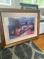 Framed print showing a colorful landscape with a rustic-style gold frame placed on a rug indoors.