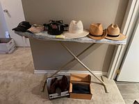 Full view of the ironing board surface displaying hats: one black Smith & Wesson cap, beige cap with H logo, white-beige straw hat, natural tan straw hat, light brown fedora. Below the board are brown Justin boots and a box with Mudruckers shoes and glove.