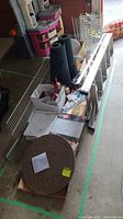 Wide view of the lot showing round floor tiles, rolls of black material, shelving units, ladder, boxes with miscellaneous items.