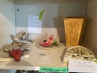 Lot of ceramics on white shelf including 2 winged heart wall plaques, a figurine on plate, and a textured yellow vase. Tagged as 'Ceramics - one lot' with note identifying limited edition tile by Grace Nickel.