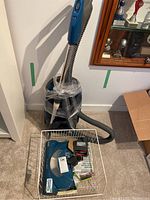 Full view of vacuum cleaner wrapped in plastic with hose, wand, and basket of accessories on carpeted floor near cabinet.