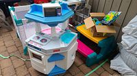 Photo showing Fisher-Price blue and white play kitchen next to Little Tikes picnic table with box of toys on top.