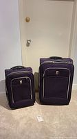 Two matching purple fabric London Fog luggage pieces side by side, taller approx 26 inches on the right, shorter on the left, shown standing upright on carpeted floor.