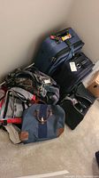 Wide angle showing lot of luggage and bags stacked, including blue wheeled suitcase, camouflage backpack, and Polo Ralph Lauren tote.