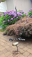 Photo showing full bird sculpture next to garden plants, highlighting stone body and metal legs and tail.